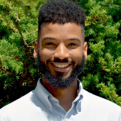Outdoor portrait of Naim Edwards standing in front of green evergreen shrubs.